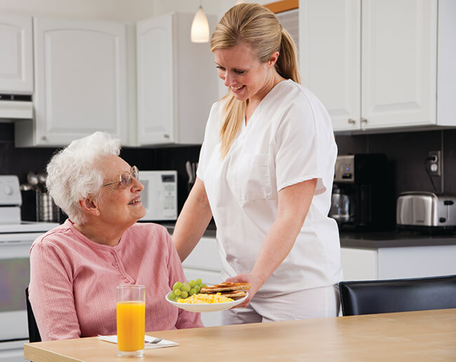 Nurse in white scrubs serving a meal to an older woman in the kitchen in Dallas, TX