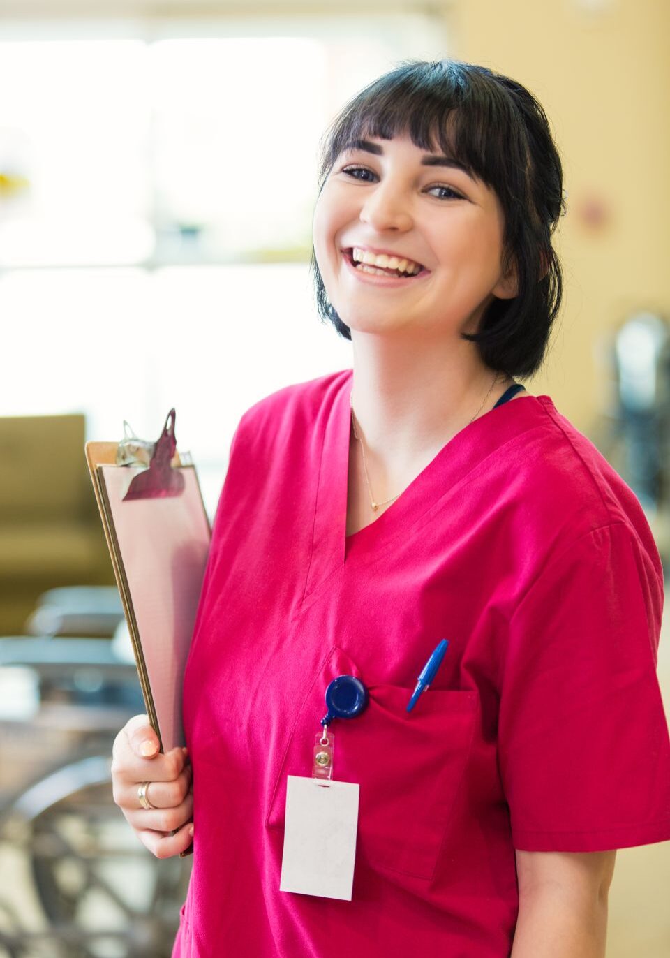 Pretty young Caucasian female nurse smiles at the camera in hospital, nursing home or rehab facility. she is holding a clipboard containing a patient's medical chart. She is also wearing an id lanyard.